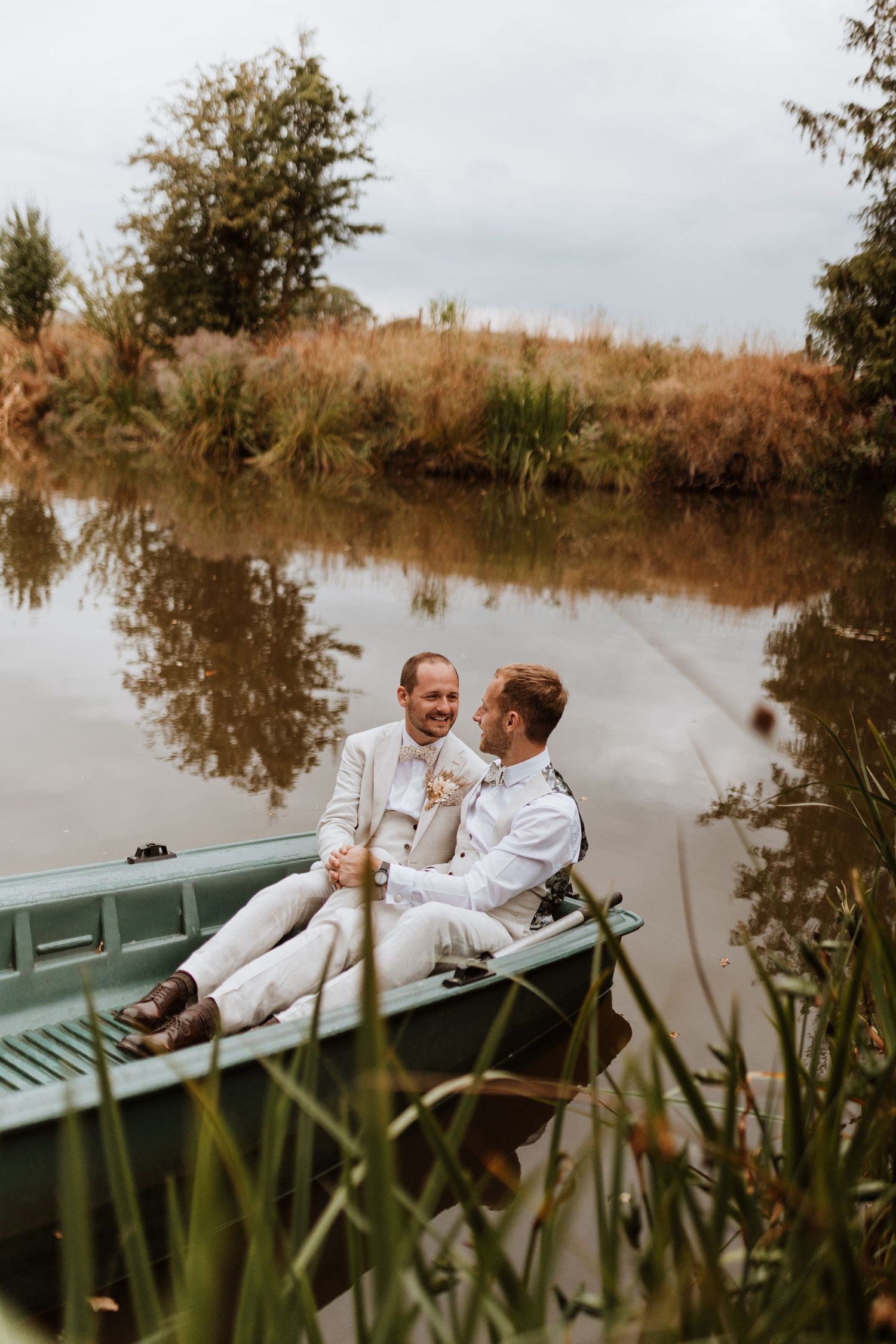 Two grooms in white suits share a romantic moment in a green row boat on a calm reflective lake on their wedding day at Moat Hall Barns