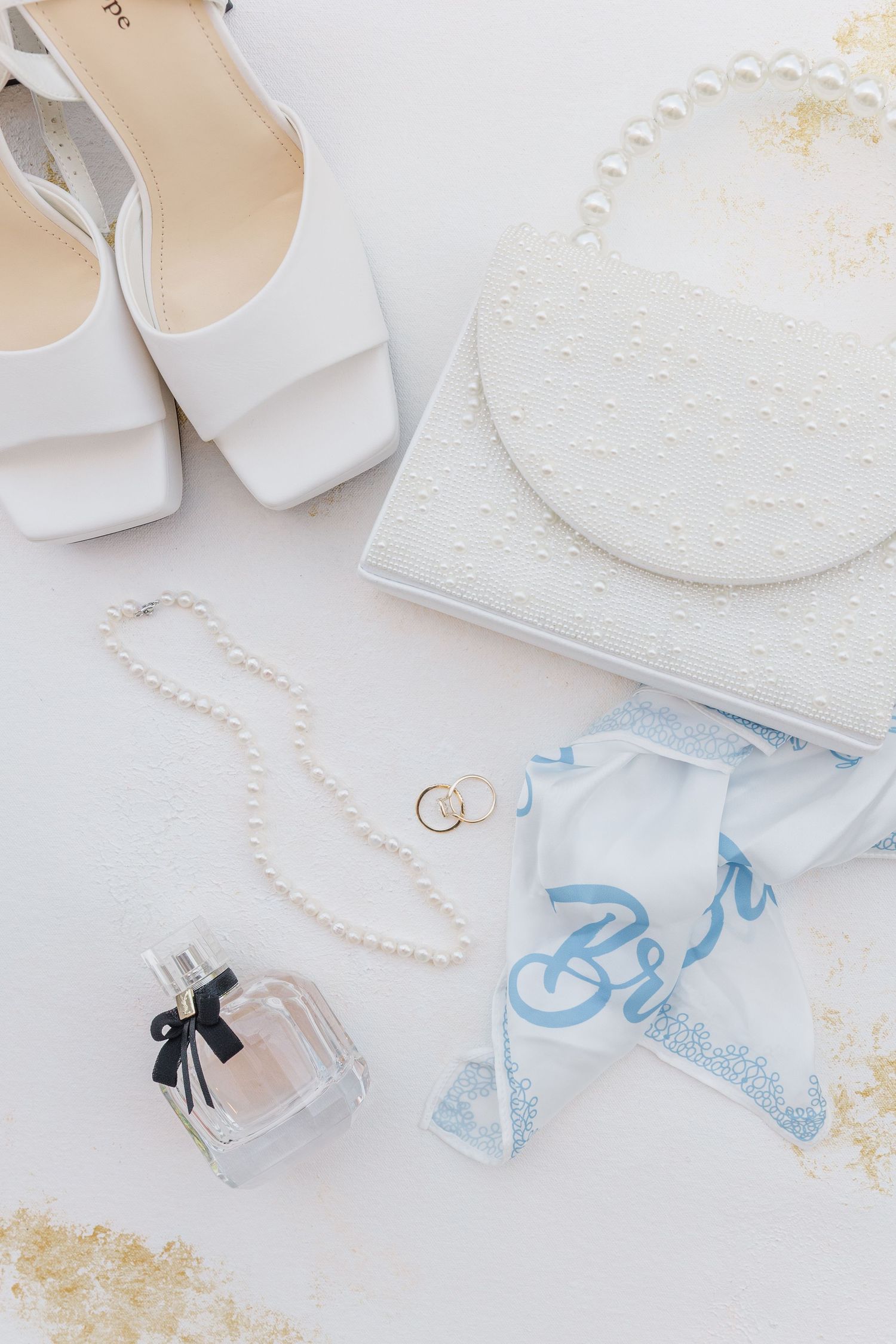 Bridal flat lay with white heels, pearl clutch, pearl necklace, gold rings, perfume, and blue bride handkerchief.