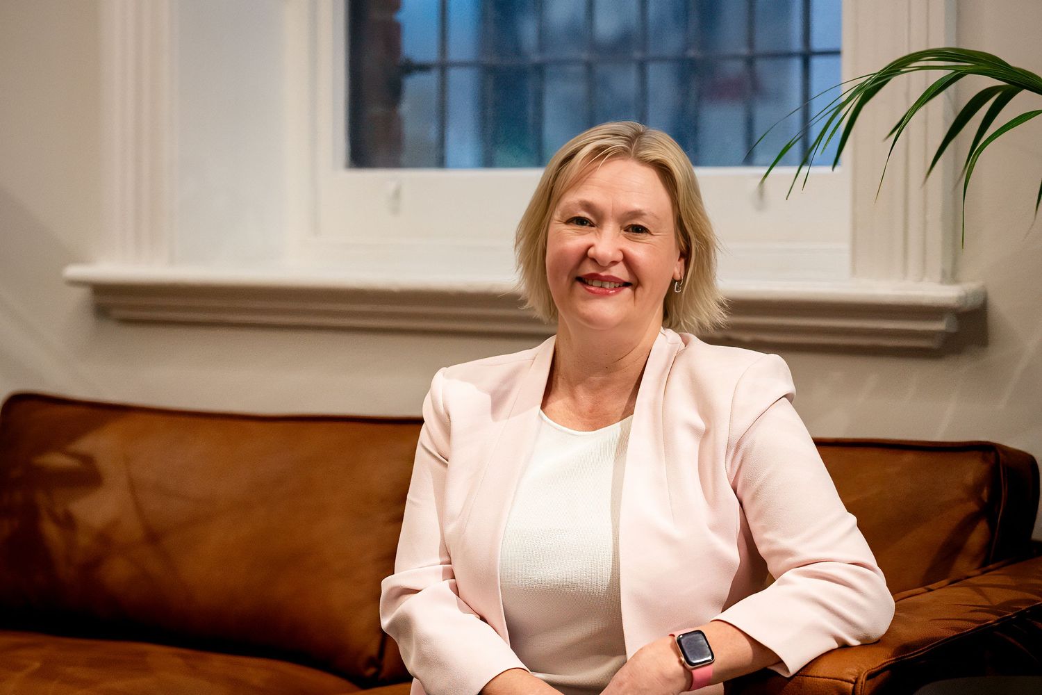 Professional woman in white blazer smiling warmly while seated on a brown leather sofa in a bright office.