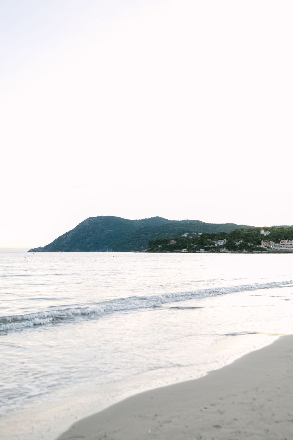 Serene beach scene with calm waves, white sand, and a lush green mountain peninsula under a bright sky.
