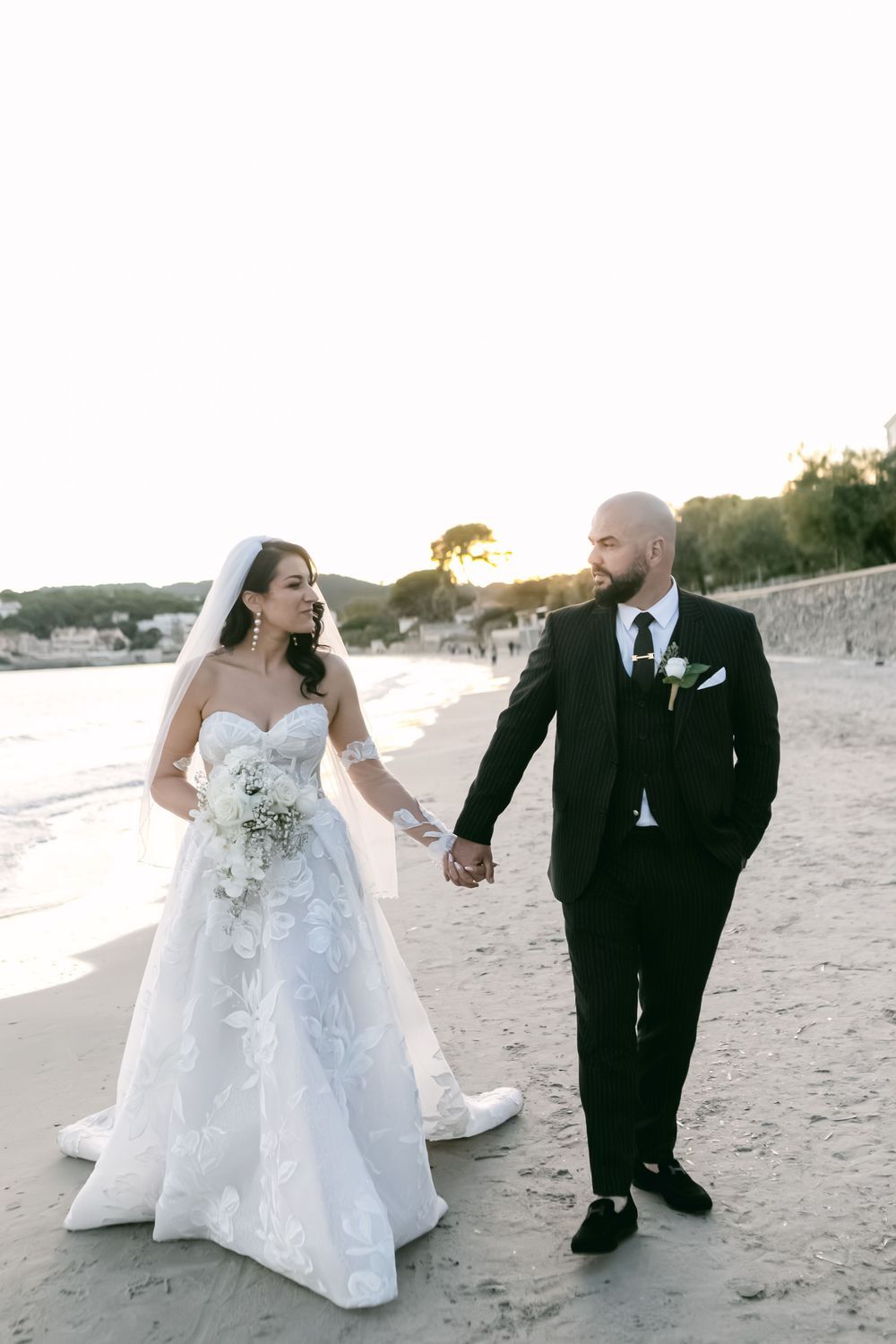 Bride and groom walk hand in hand on a sandy beach at sunset, bride in white lace gown with veil and bouquet.