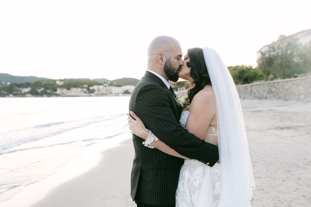 Bride and groom sharing a romantic kiss on a sandy beach at sunset, bride wearing a white gown and veil.