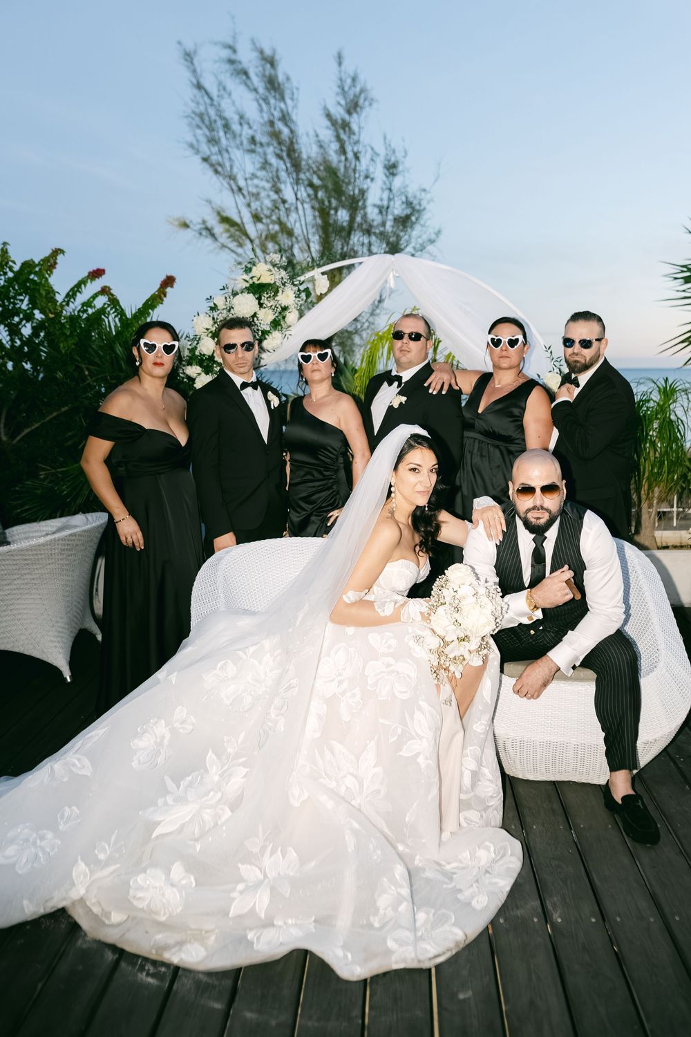 Wedding party in black attire with sunglasses poses with bride in white gown and groom on a deck by the ocean.