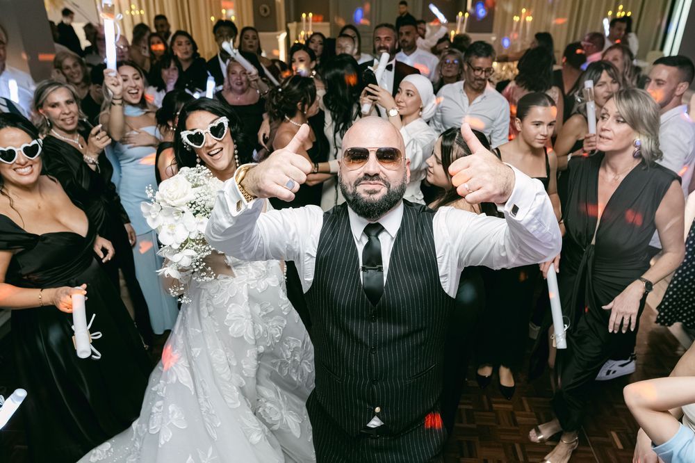 Joyful bride and groom giving thumbs up surrounded by celebrating wedding guests on the dance floor.