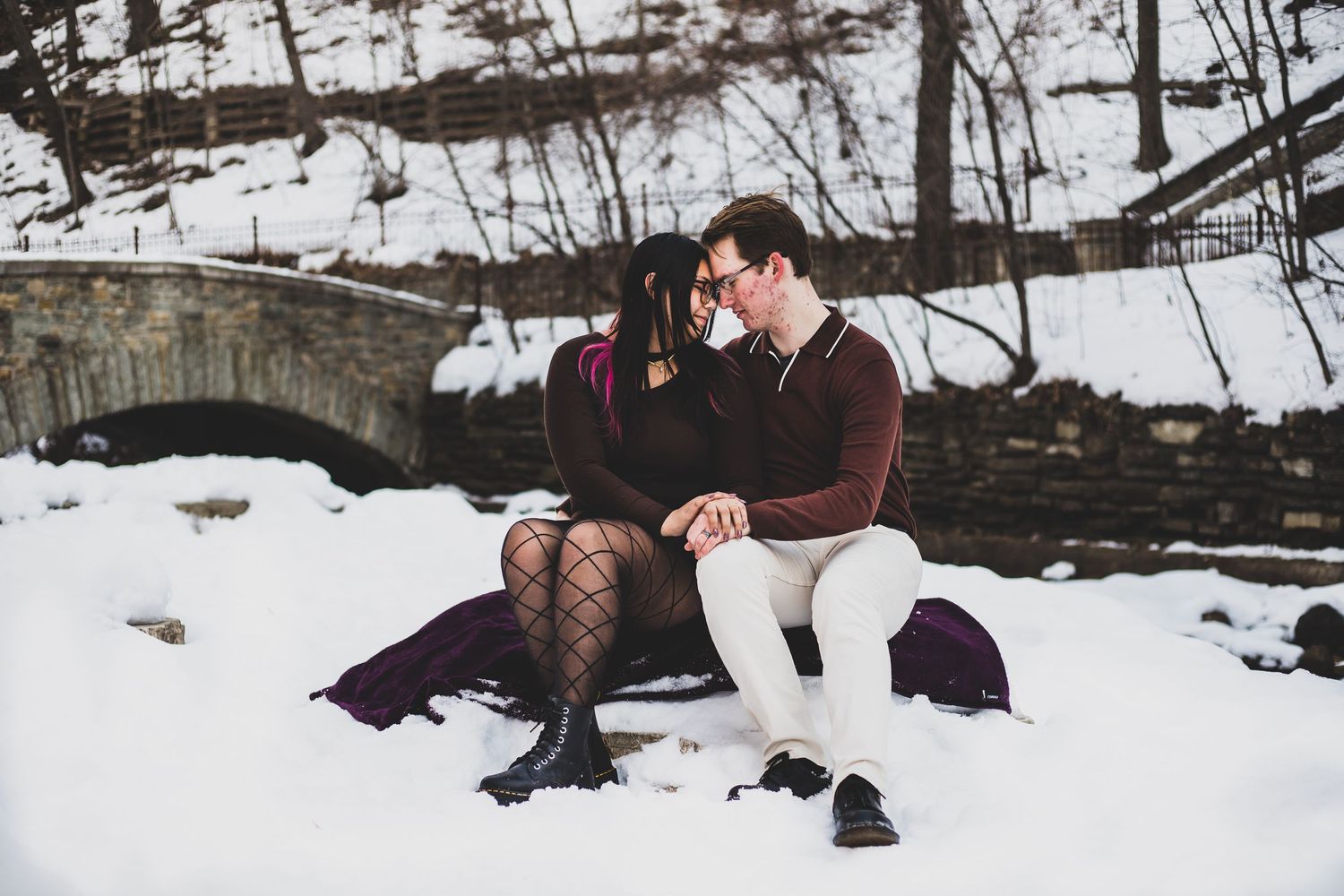 Couple relaxing together on snow-covered ground near stone bridge in winter setting.