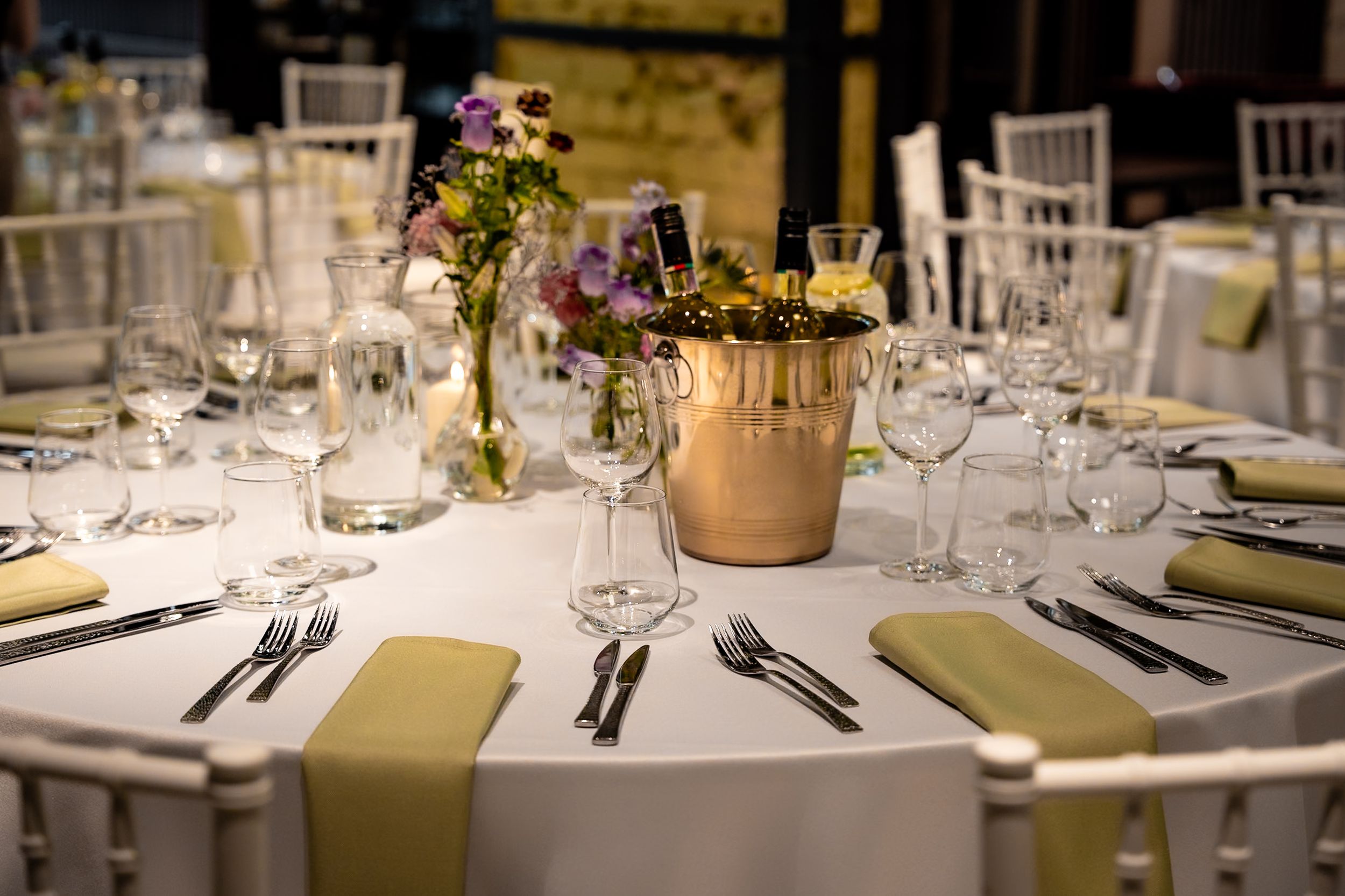 Elegant wedding reception table set with gold centerpiece, floral arrangement, crystal glassware, and olive green napkins.
