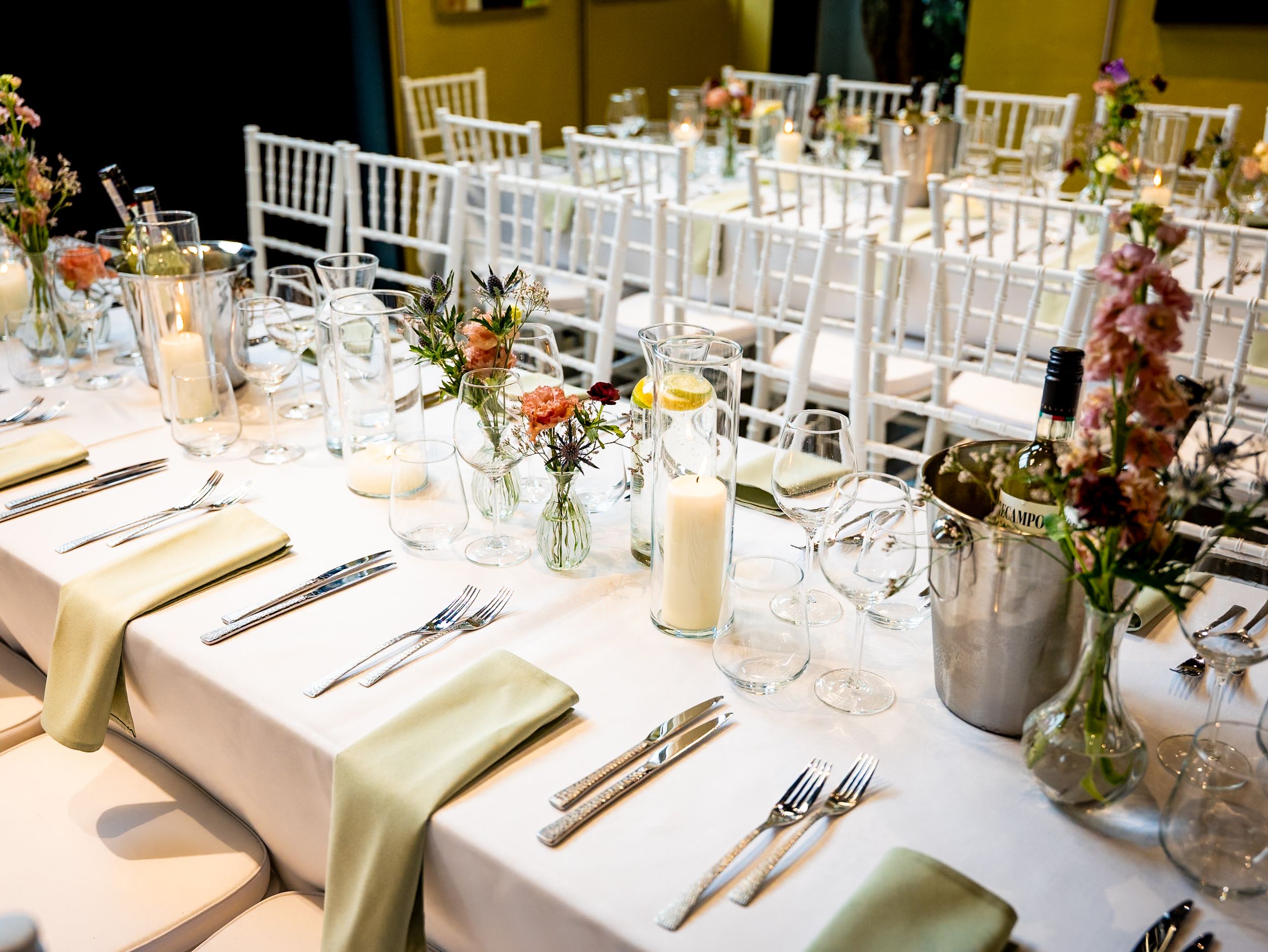 Elegant wedding reception table set with white linens, candles, floral centerpieces, and glassware in a bright venue.