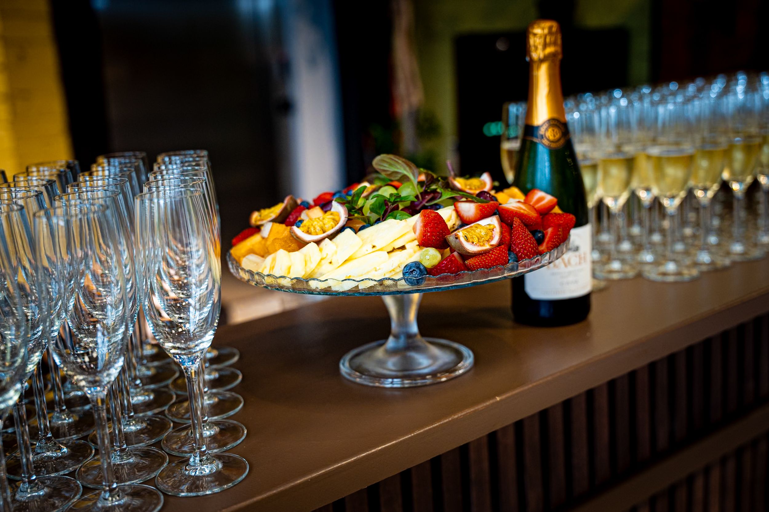 Elegant champagne flutes and a fruit platter with cheese displayed on a wooden bar beside a champagne bottle.