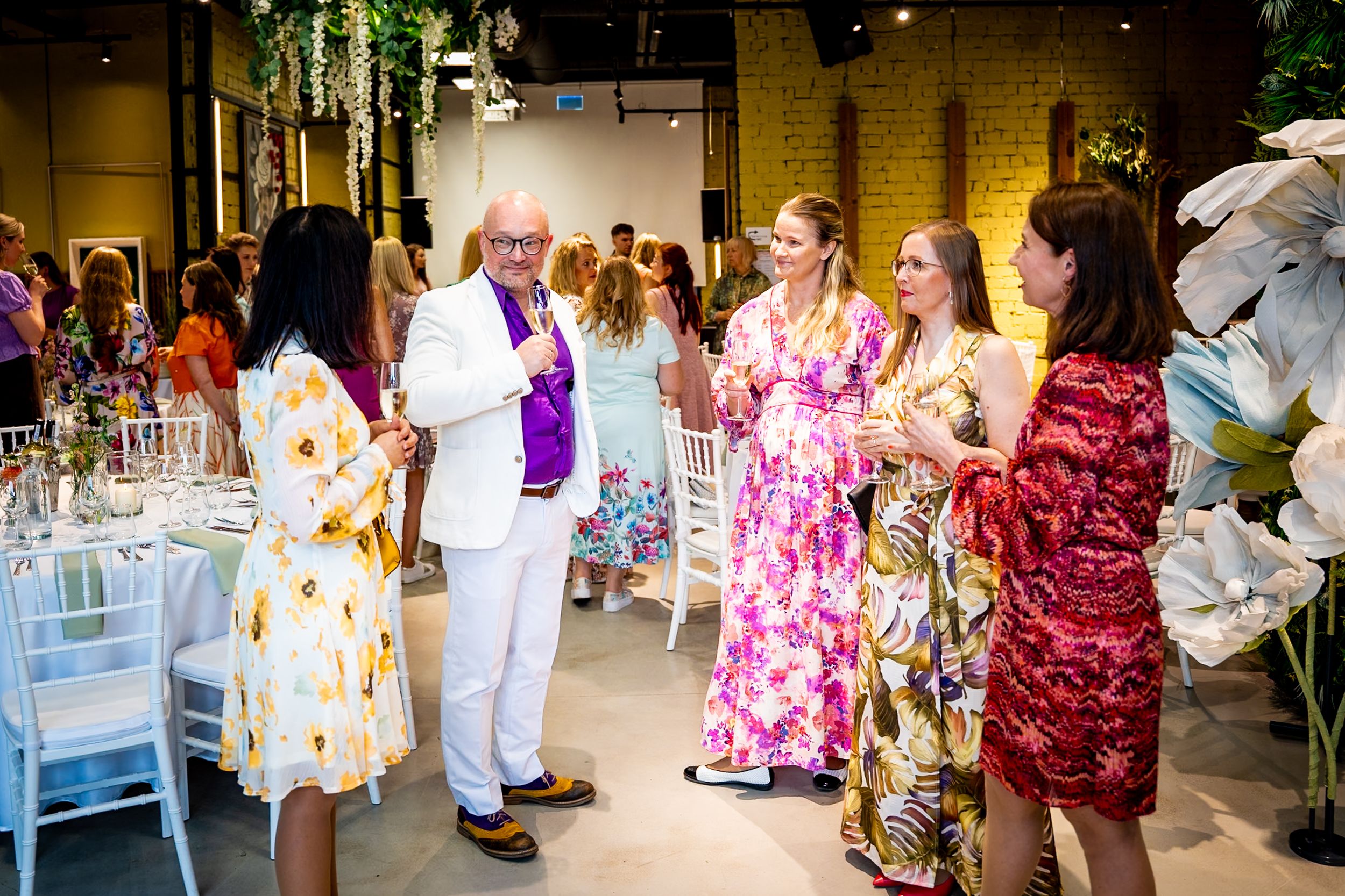 Elegantly dressed guests mingle at a colorful indoor event, wearing floral gowns and white suit in a decorated venue.