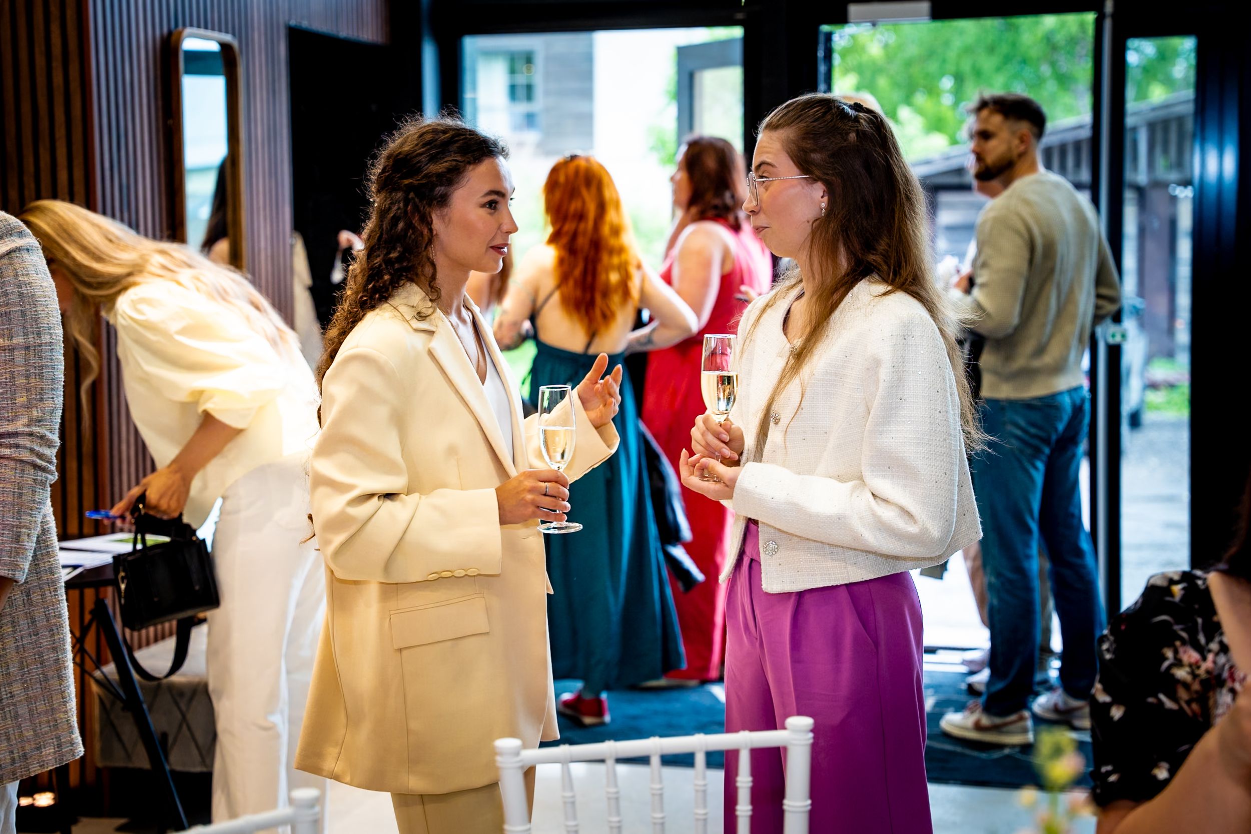 Two women in elegant attire chat at a sophisticated social gathering, holding drinks in a bright modern venue.
