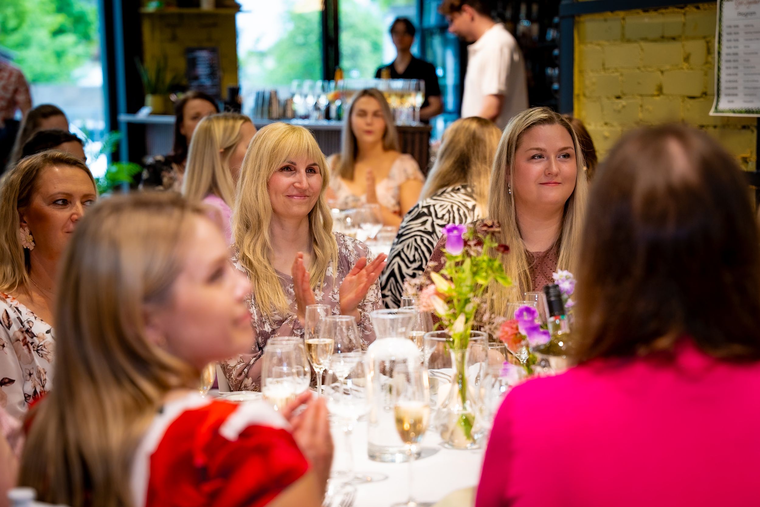 Women gathered at an elegant dining event, smiling and clapping around a flower-adorned table in a bright restaurant.