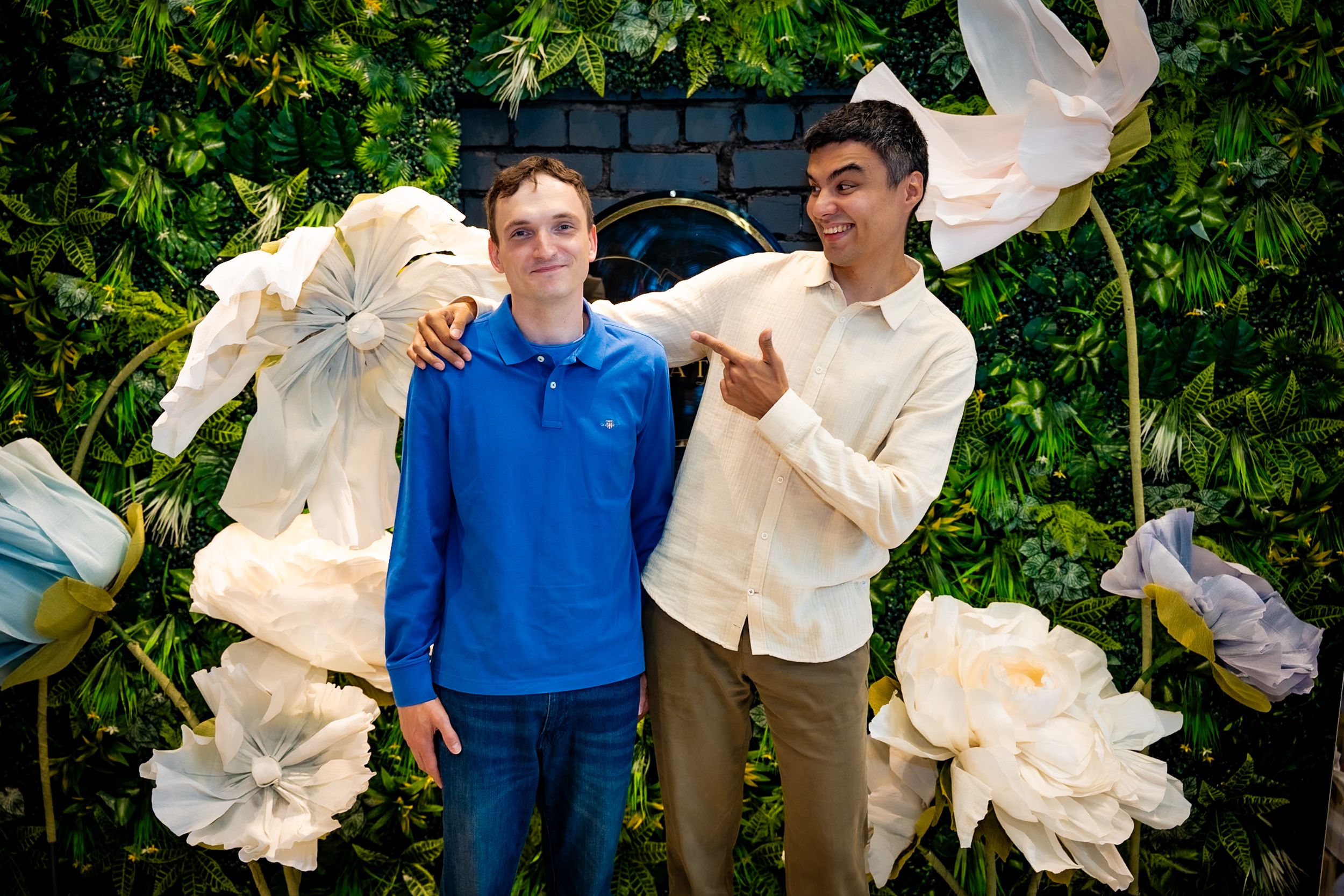 Two young men smiling and posing together in front of a decorative green wall with large white paper flowers.