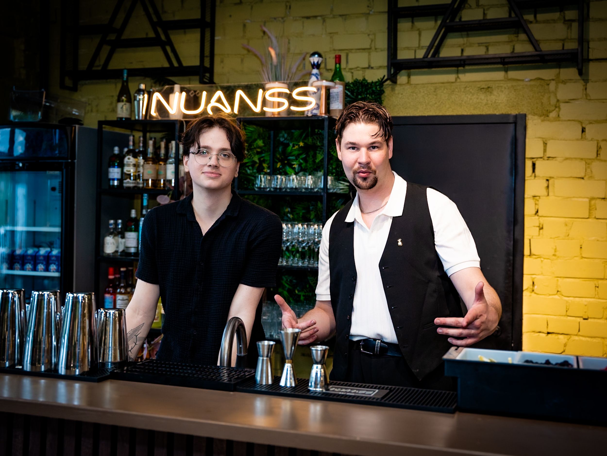Two bartenders standing behind a bar at Nuanss cocktail lounge, surrounded by bottles and shakers.
