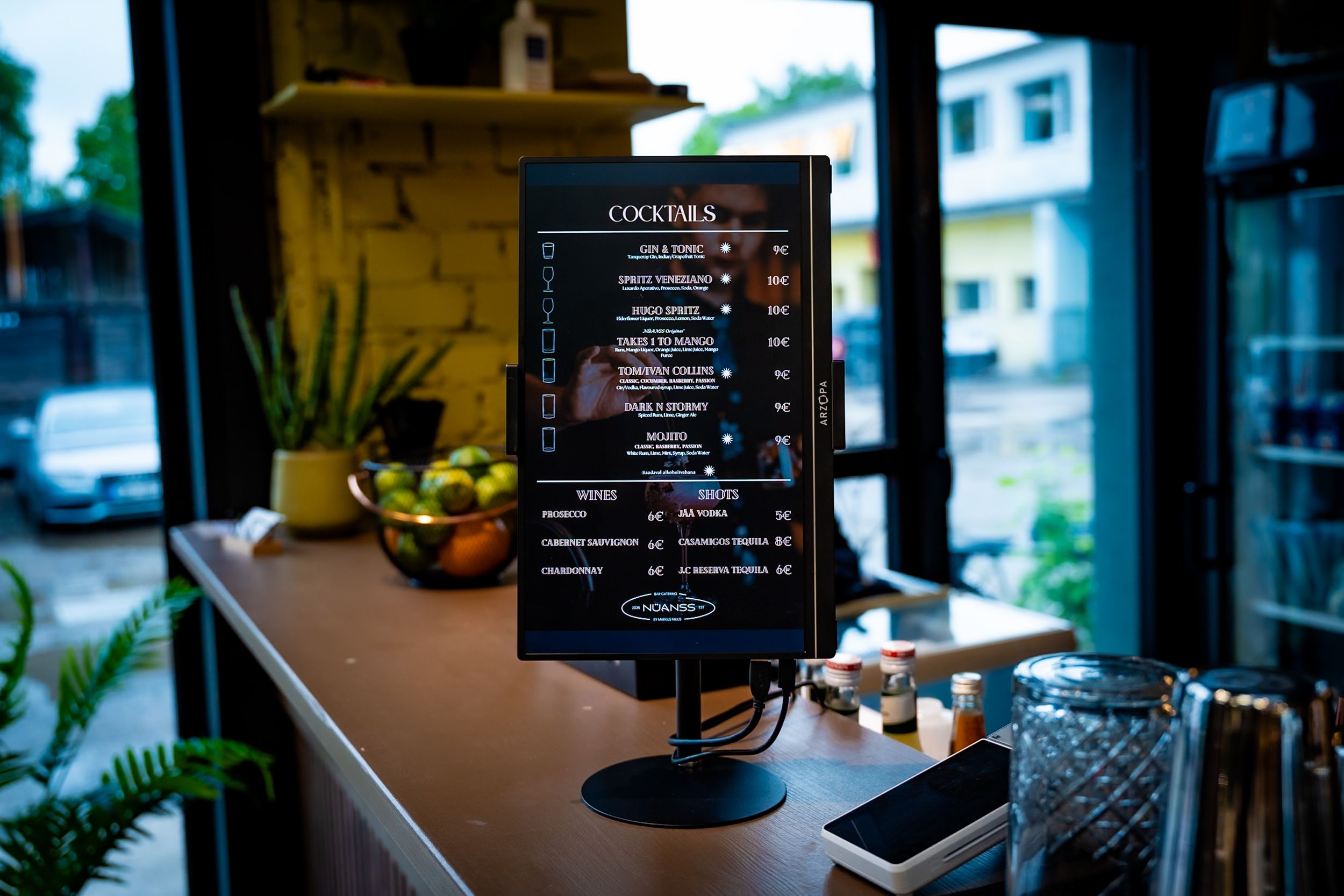 A digital cocktail menu display on a bar counter near a window in a dimly lit restaurant.