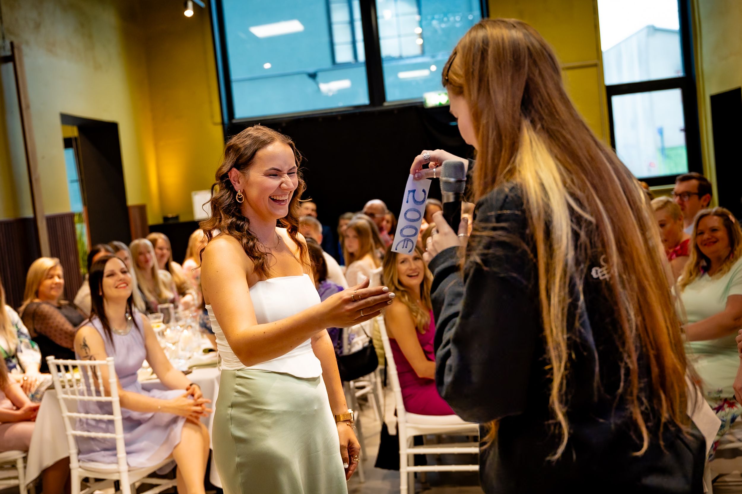 Woman in white dress receives gift from long-haired girl at elegant indoor celebration with seated guests.