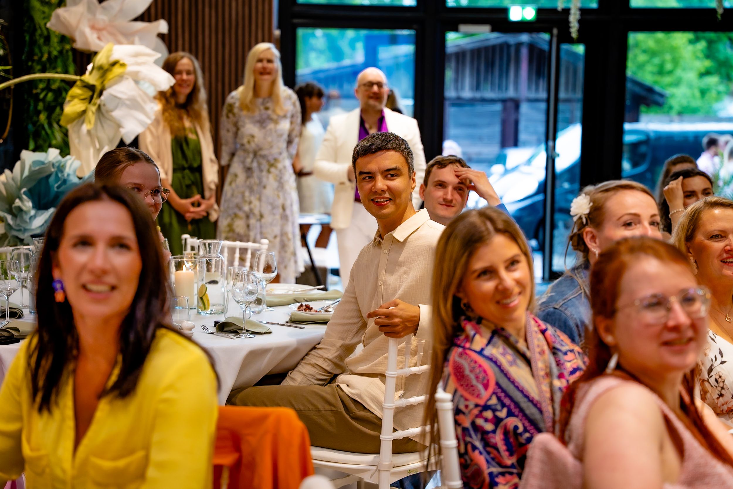 Guests smiling and laughing at an elegant indoor wedding reception with floral decorations and white chairs.