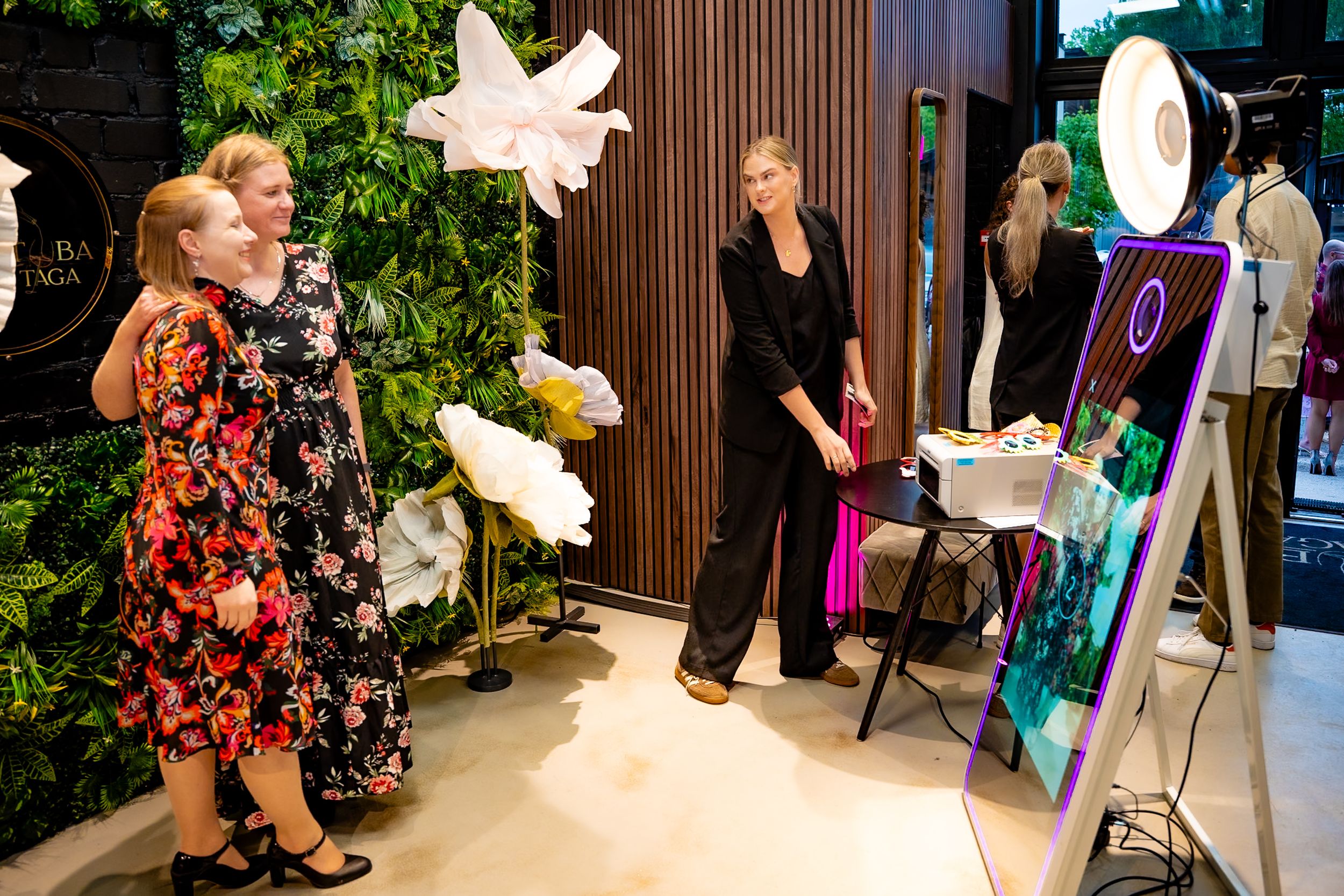Two women pose at a mirror photo booth with floral wall backdrop at an elegant indoor event.