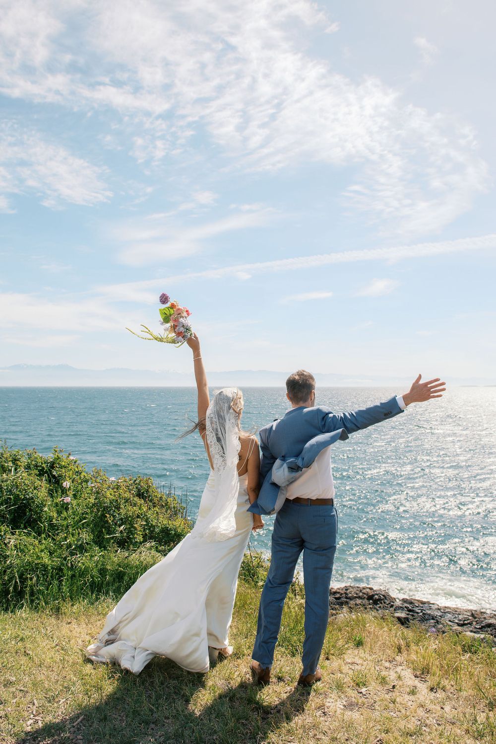 Bride and groom celebrate on coastal cliff, arms raised, holding bouquet, wearing wedding dress and blue suit, ocean backdrop.
