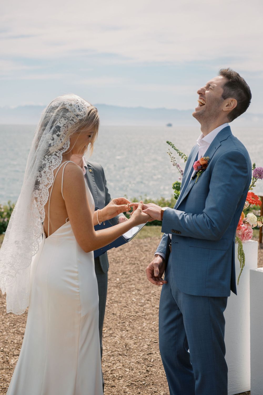 Bride placing ring on groom's finger as he laughs joyfully during outdoor wedding ceremony by the ocean.