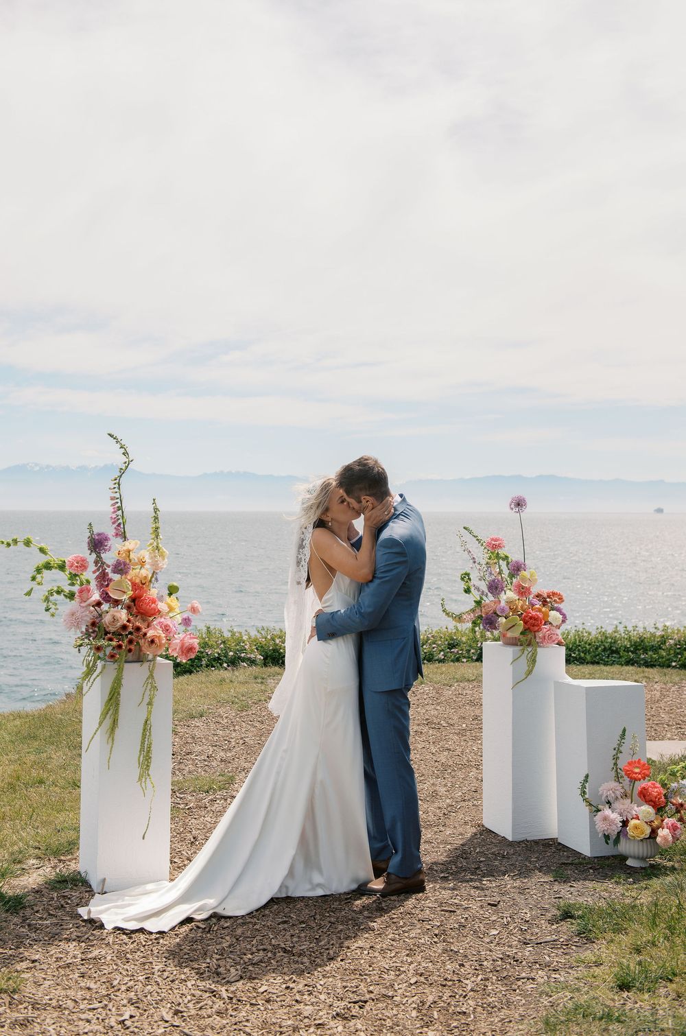Bride and groom share first kiss at outdoor waterfront wedding ceremony flanked by colorful floral arrangements on white pedestals.