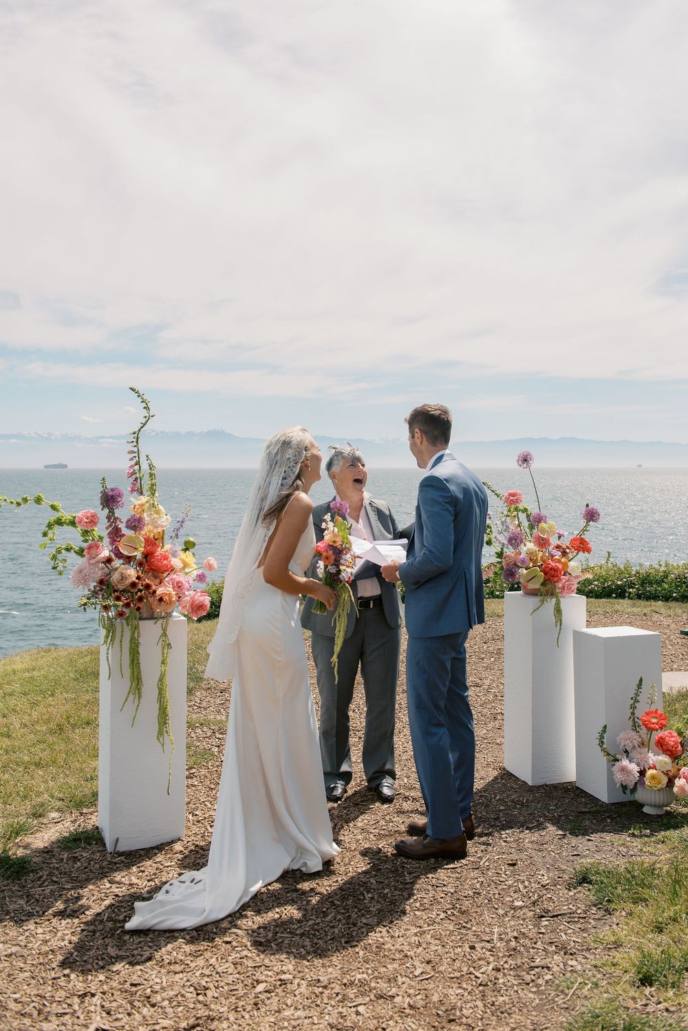 Outdoor wedding ceremony by the ocean with bride and groom exchanging vows, flanked by colorful floral arrangements on white pedestals.