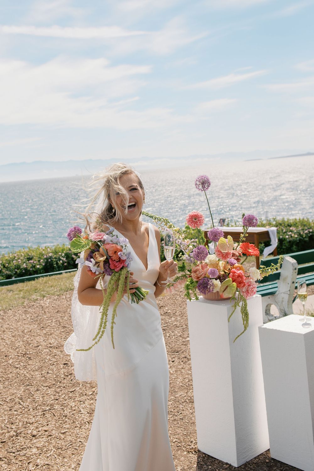 Smiling bride in white gown holds colorful bouquet outdoors near ocean with vibrant floral arrangements on white pedestals.