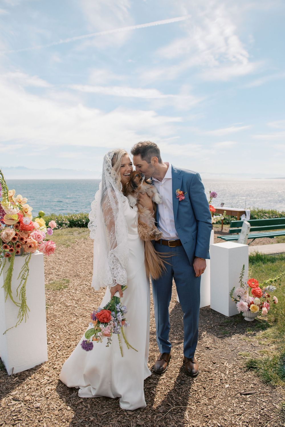 Bride and groom holding their dog at an outdoor coastal wedding ceremony with colorful floral arrangements.
