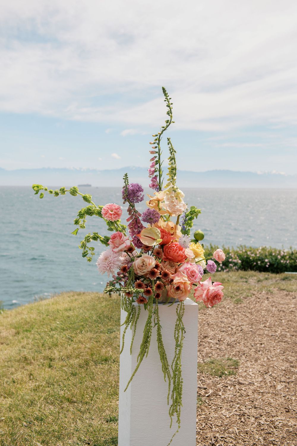 Colorful floral arrangement on white pedestal with ocean backdrop featuring pink roses, foxglove, and mixed blooms.