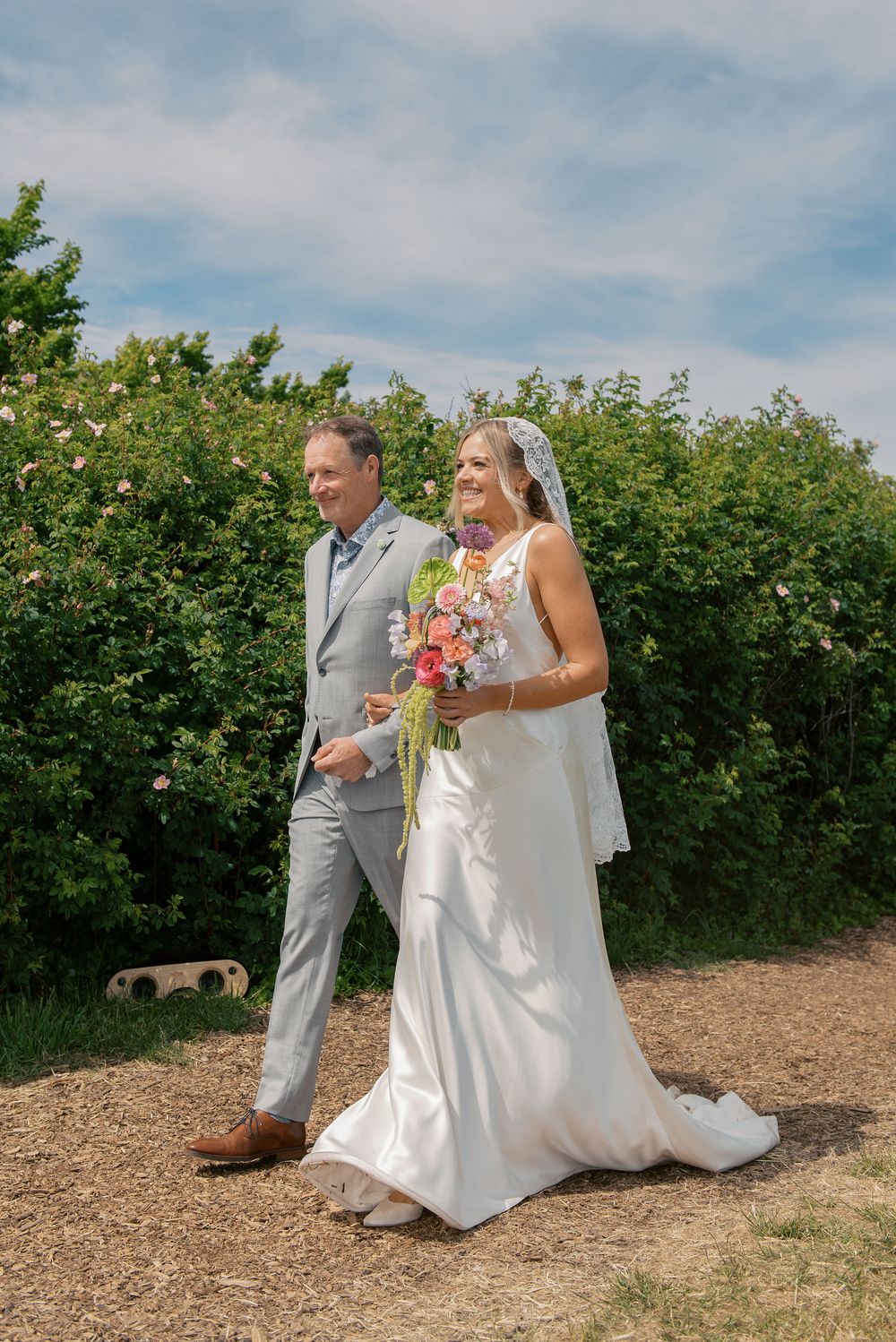 Bride in white gown holding colorful bouquet walks with man in gray suit outdoors on sunny wedding day.