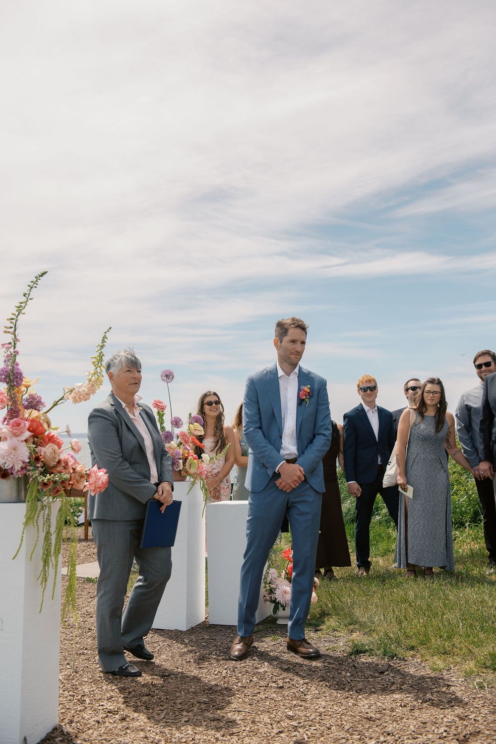 Groom in blue suit stands at outdoor wedding altar with colorful floral arrangements, officiant, and guests under blue sky.