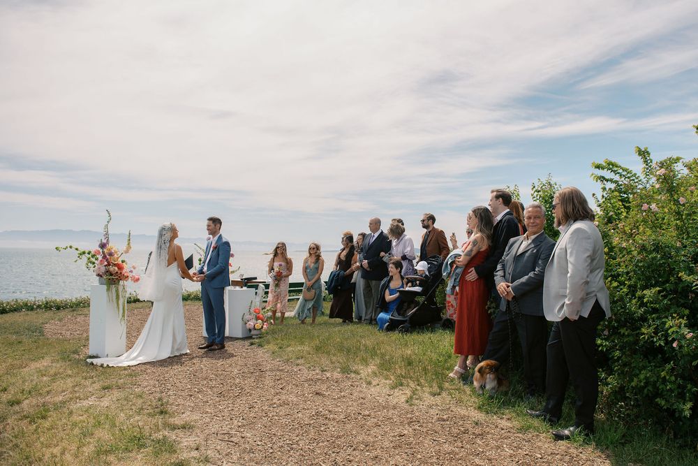 Outdoor wedding ceremony on a coastal cliff with bride and groom at altar, guests standing nearby under a partly cloudy sky.