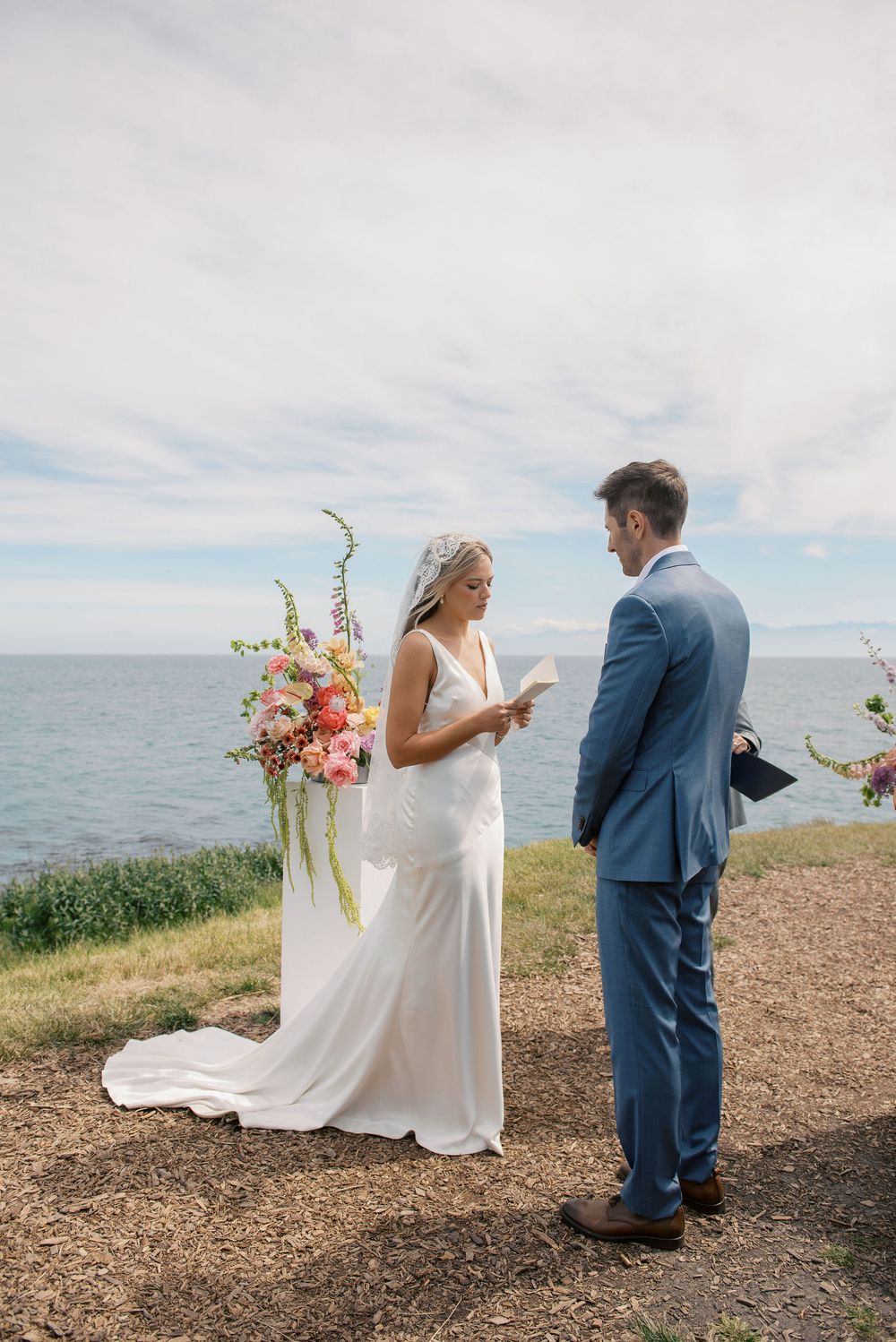 Bride and groom exchange vows outdoors by the ocean, with colorful floral arrangement, bride in white gown, groom in blue suit.