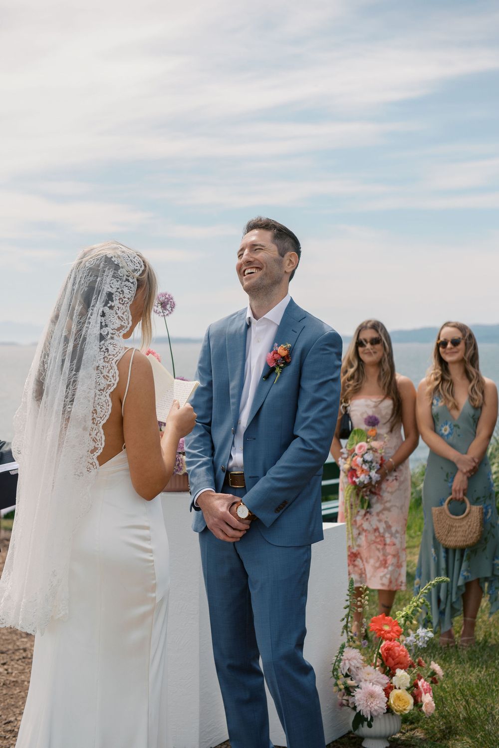 Groom in blue suit laughing joyfully at bride during outdoor wedding ceremony by the ocean with bridesmaids.
