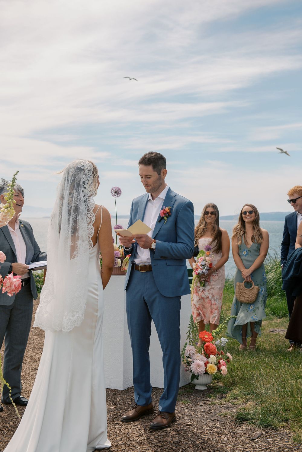 Bride and groom exchanging vows outdoors by the ocean, surrounded by guests at a sunny seaside wedding ceremony.