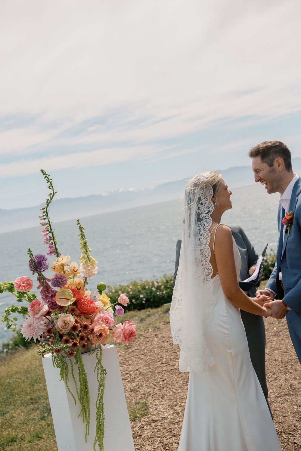 Bride and groom holding hands at outdoor wedding ceremony with colorful floral arrangement and ocean backdrop.