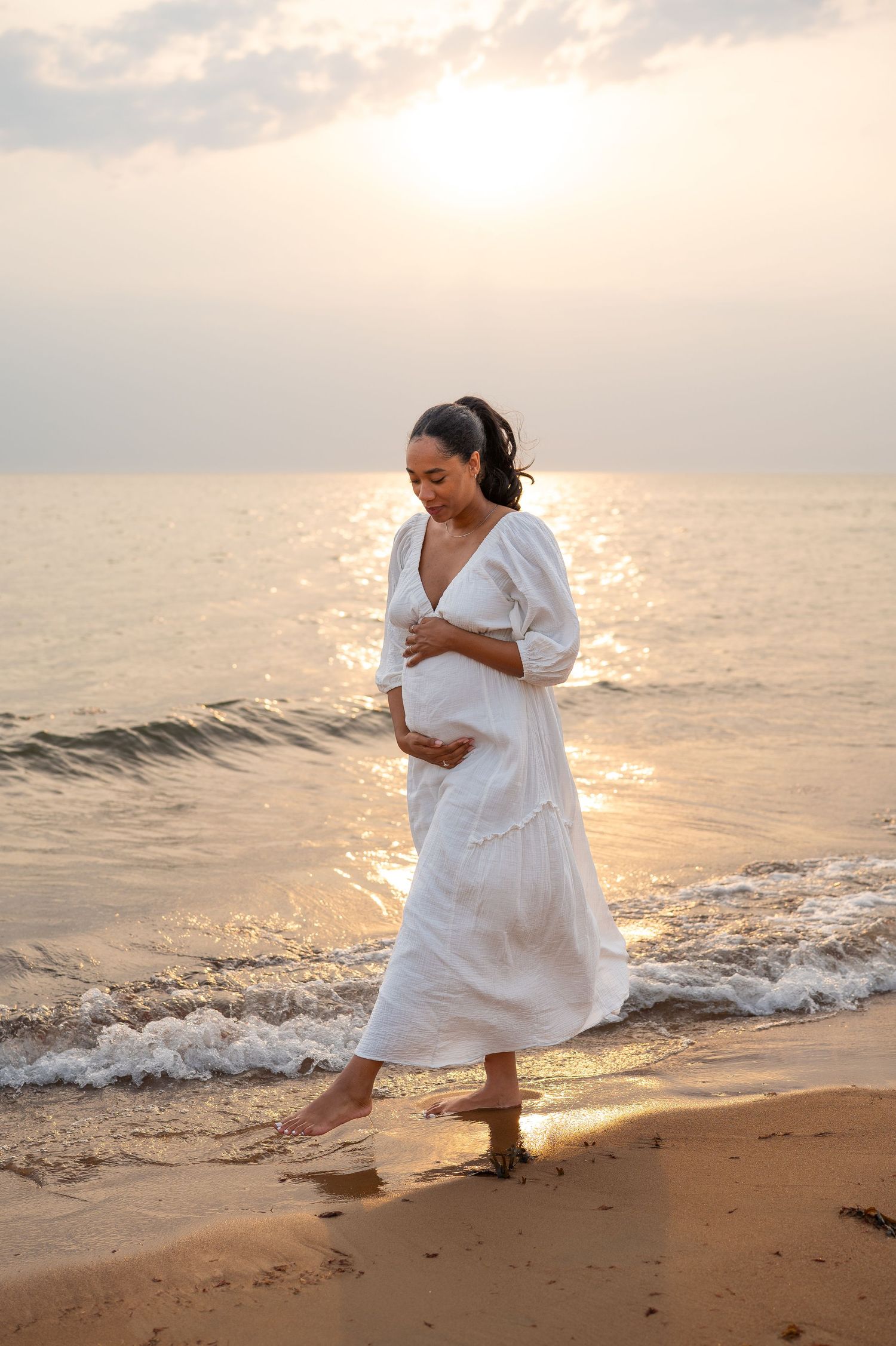 Pregnant woman in white dress walking on beach at sunset.