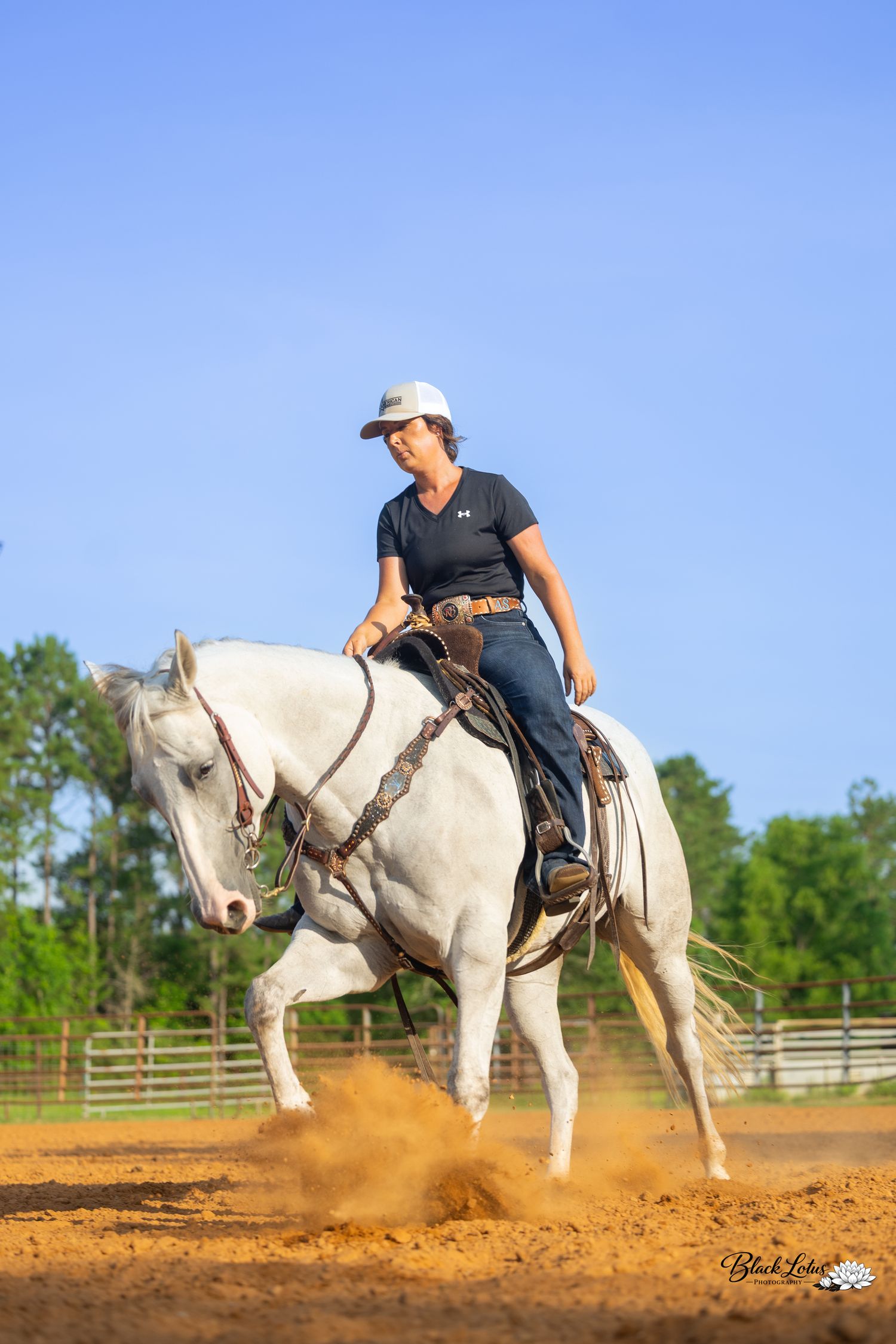 A rider in a white hat and navy shirt performs a reining maneuver on a white horse in a sandy arena.