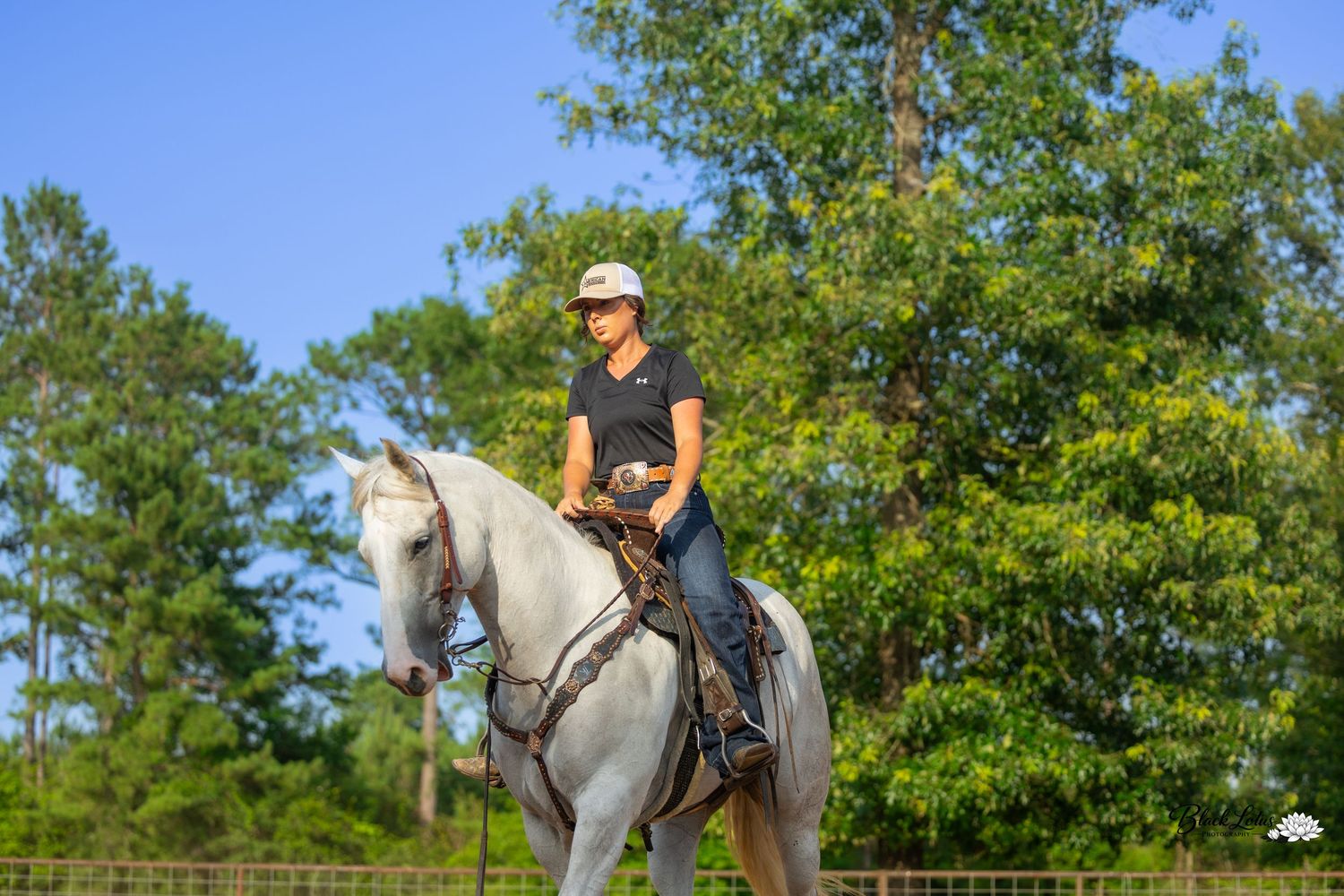 A woman in a white cap rides a white horse in an outdoor arena with lush green trees in the background.