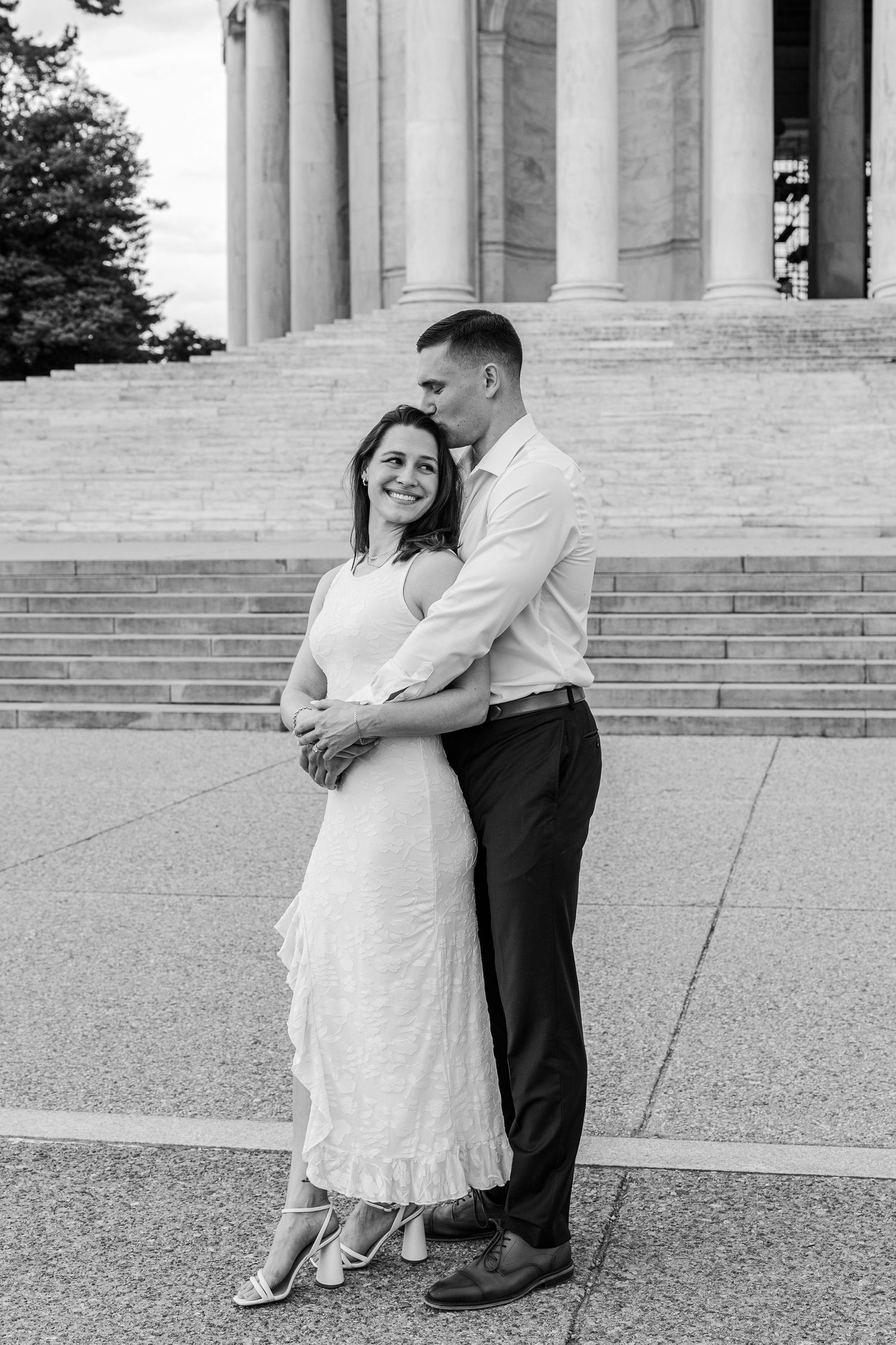Black and white photo of a couple embracing on steps of a neoclassical building, woman in white dress smiling at camera.