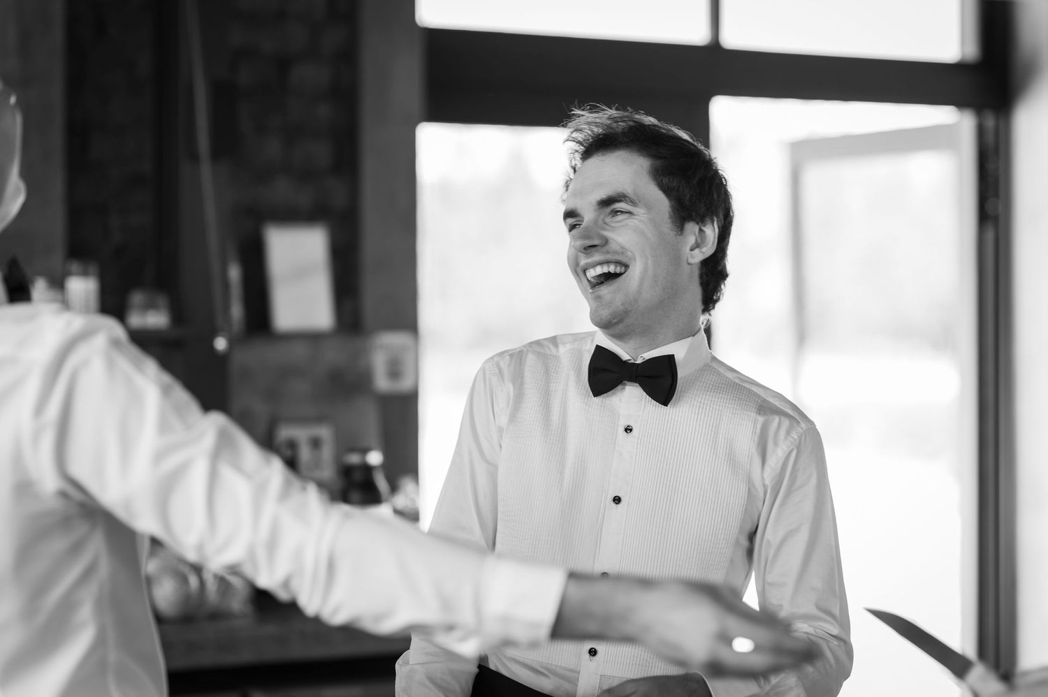 Smiling groom in white dress shirt and black bow tie laughing joyfully at a wedding reception.