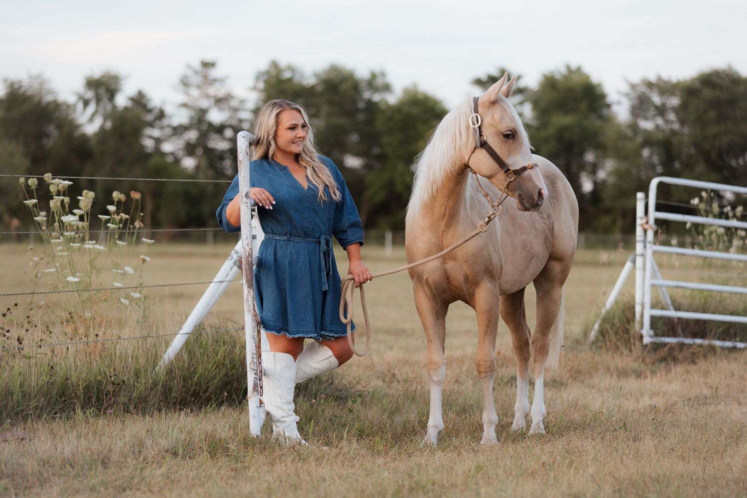 Woman in blue dress and white boots stands beside a palomino horse in a pastoral field with trees.