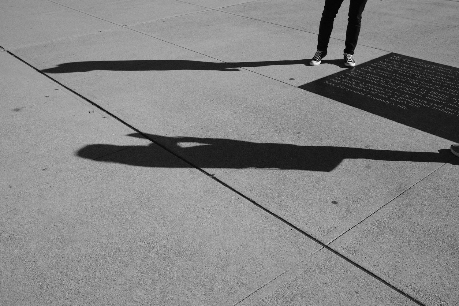 Black and white photo of a person's legs casting a long dramatic shadow on a concrete sidewalk.