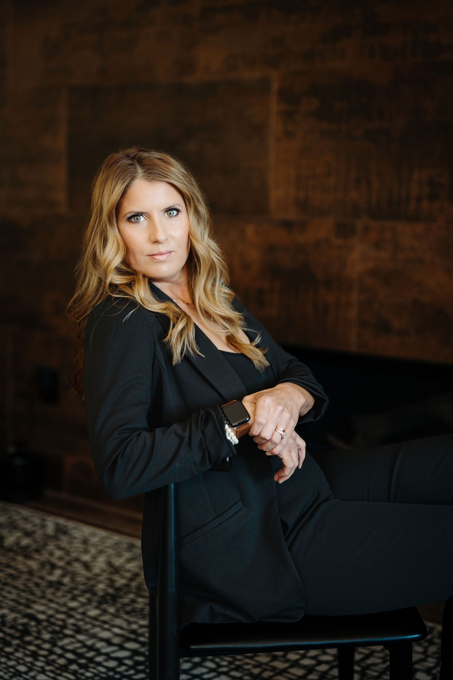 Woman in black suit sitting confidently in chair against dark brick wall backdrop.