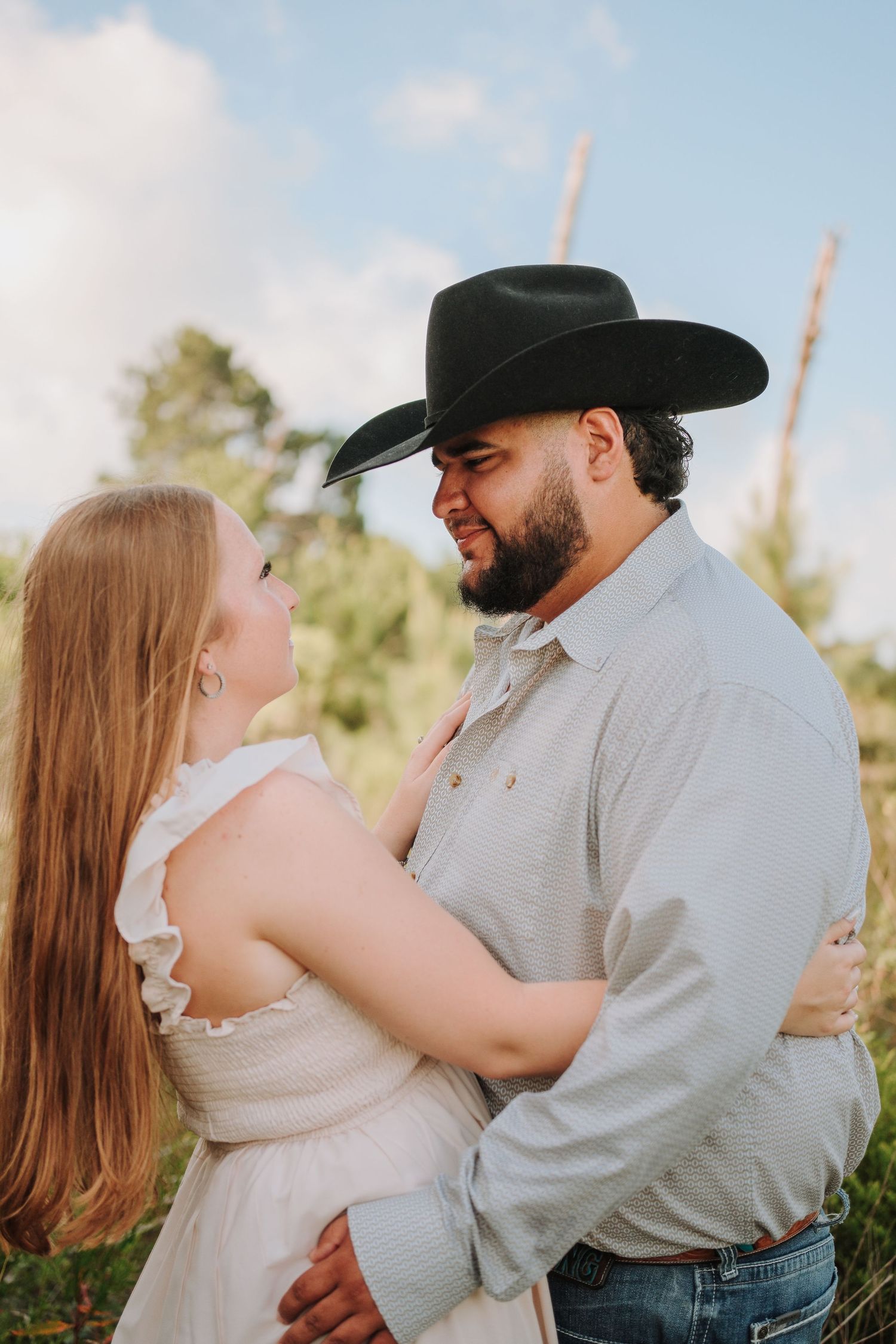 A couple in a field, woman in white dress, man in black cowboy hat, sharing a romantic embrace outdoors.