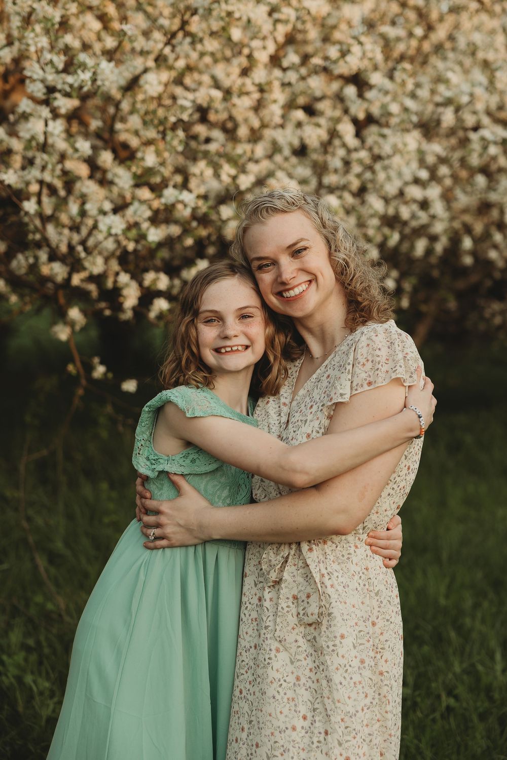 A smiling woman and young girl in floral and mint dresses embrace warmly in front of blooming white spring flowers.