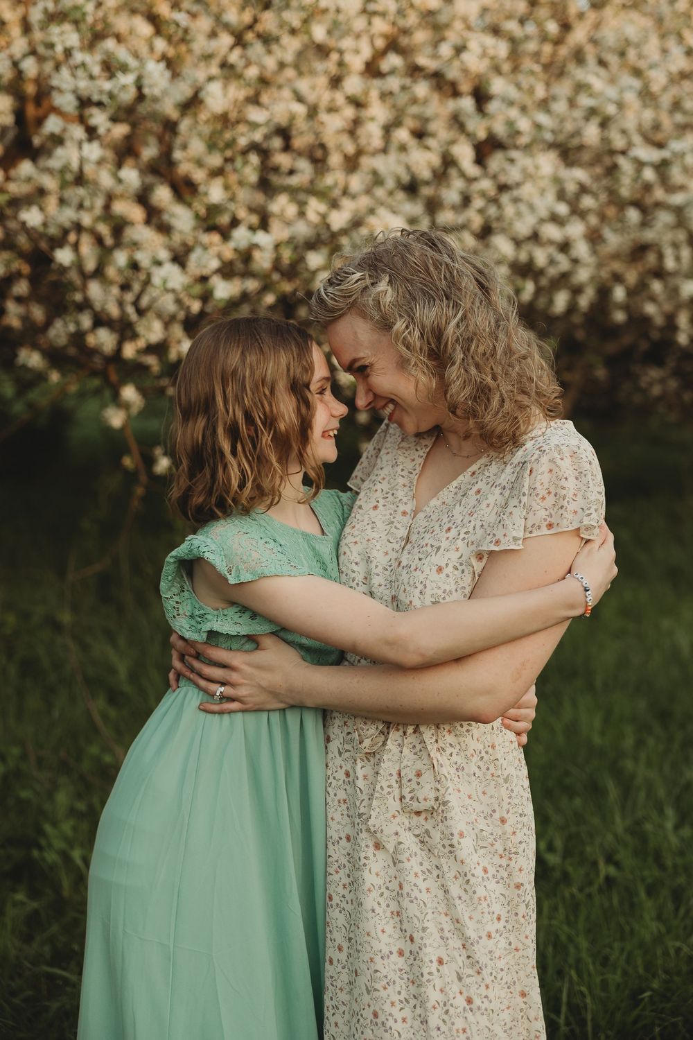 Two women in floral and mint dresses embrace warmly in front of a blooming white flower tree outdoors.