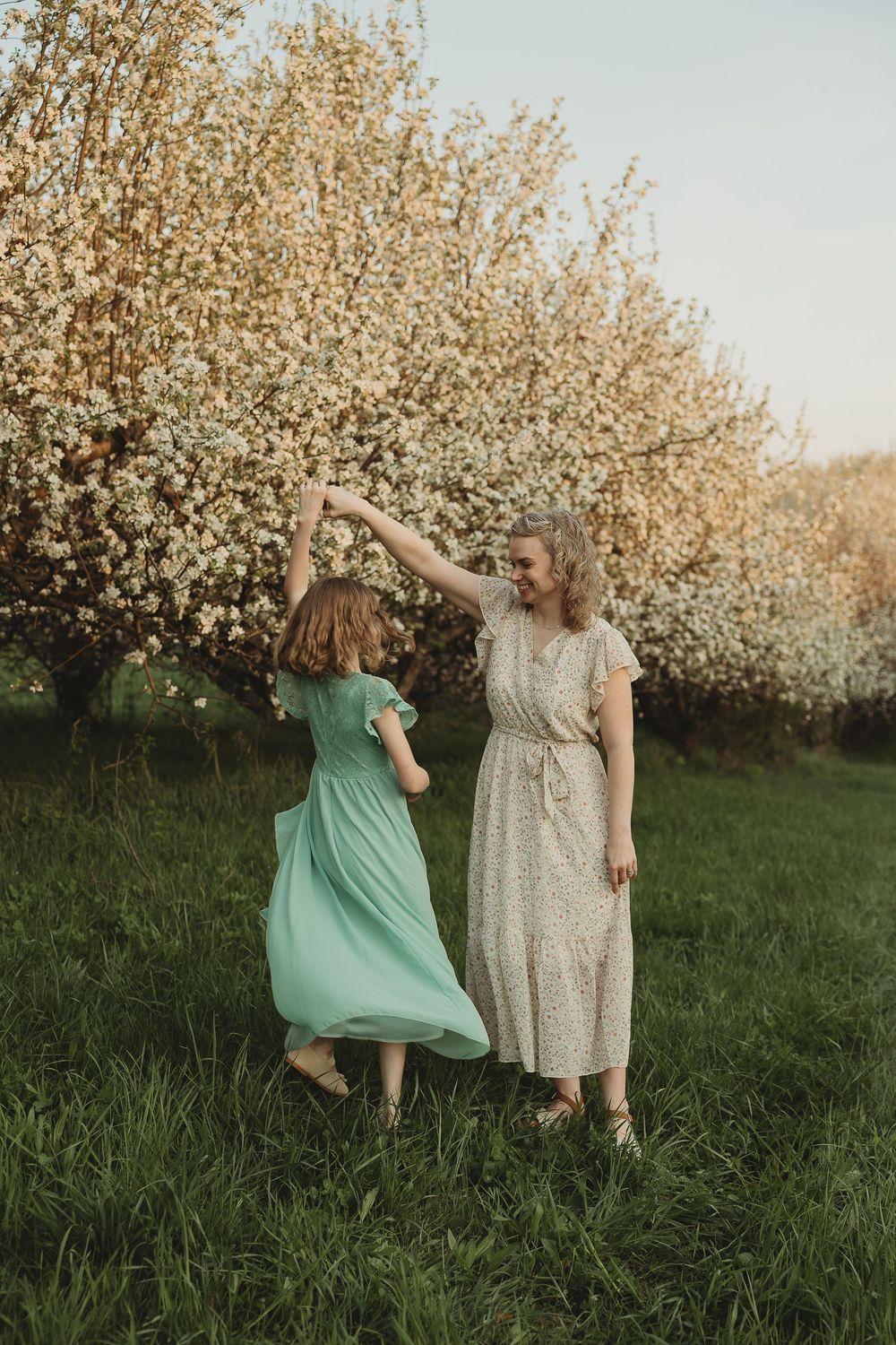 Two women in floral dresses dancing joyfully in a blooming spring orchard on a lush green lawn.
