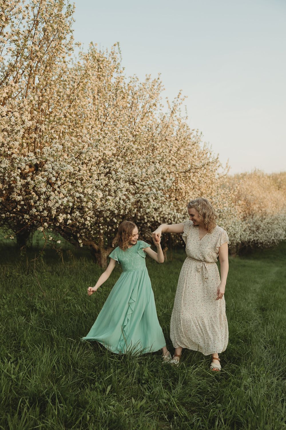 Two women in elegant dresses dancing in a blooming orchard during golden hour.