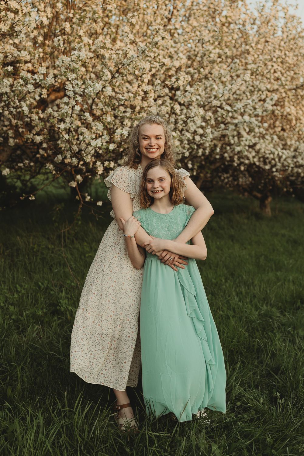 Mother and daughter in floral and mint dresses embrace in a blooming spring garden with white flowering trees.