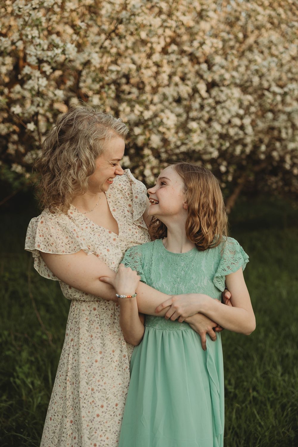 A woman and young girl in floral and mint dresses laugh together in front of blooming white flowers.